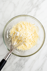 A top-down view of a large clear glass mixing bowl placed on a white marble countertop, filled with 1/2 cup cream cheese, 1/2 cup shredded Double Gloucester cheese, 2 tablespoons of heavy cream, and 1/4 teaspoon of coarse salt. A stainless steel whisk with a black handle is partially mixing the ingredients, showing the start of the creamy, flavorful base for the sandwich. Each ingredient is clearly visible, and the scene is simple, clean, and focused on preparation.