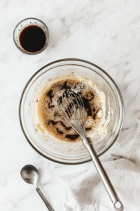 In this image, a large clear glass mixing bowl sits on a white marble countertop. Inside, eggs, light and dark corn syrups, white sugar, melted butter, vanilla extract, and salt are being whisked together until smooth. This well-blended filling is the rich and sweet heart of the mini pecan pie recipe, ready for use.