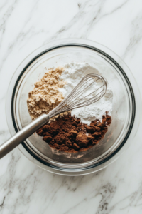 All-purpose flour, baking powder, salt, and sugar are being combined in a clear glass mixing bowl on a white marble countertop. A whisk is resting inside the bowl, ready to blend the dry ingredients evenly. This mixture will later be combined with the carrot base to form the soufflé batter.