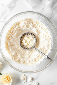 This step shows the beginning of the tart crust preparation. A clear glass bowl on a white marble countertop contains a mix of all-purpose flour, confectioners’ sugar, and salt. Small pieces of very cold butter are being cut in with a pastry cutter, forming a crumbly texture. An egg yolk is ready to be mixed in. The setup is clean and minimal, demonstrating the key early stage of tart dough formation.