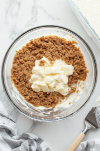 Top-down view of a clear glass mixing bowl containing graham cracker crumbs, melted unsalted butter, and granulated sugar being mixed on a white marble countertop. A metal spoon or spatula rests on the side as the ingredients are blended to form the base crust for the cheesecake. This step is essential for creating a buttery, crunchy foundation.