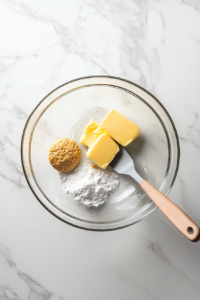 A large clear glass bowl on a white marble countertop filled with graham cracker crumbs, melted butter, and white sugar being mixed using a rubber spatula. This step ensures all ingredients are evenly moistened, forming the base of the cheesecake crust. The background is clean and minimal, focusing on the preparation of the dessert’s foundation.