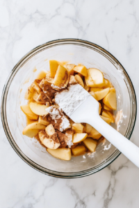Granny Smith apple slices are being mixed with white sugar, all-purpose flour, cinnamon, and nutmeg in a large glass mixing bowl set on a white marble countertop. The apples are peeled, cored, and sliced evenly to ensure a tender and flavorful filling with a perfect balance of sweet and spiced notes.