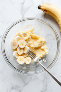 A medium glass bowl on a white marble countertop contains mashed bananas, cracked eggs, unsweetened applesauce, and skim milk. The ingredients are partially combined using a spoon. This step creates the wet base for the muffins and highlights the use of wholesome ingredients like banana and applesauce for natural moisture.