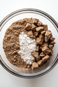 A glass mixing bowl filled with dry ingredients like medium oatmeal, all-purpose flour, baking powder, ground ginger, mixed spice, ground mace, and salt sits on a white marble countertop. Each ingredient is visible and unmixed, capturing the beginning of the recipe. The setup is clean and focused, essential for combining flavors evenly.