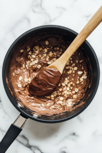 The saucepan now contains oats, creamy peanut butter, and a splash of vanilla extract atop the hot chocolate syrup. A wooden spoon begins mixing everything into a thick, chunky texture. This step blends the rich flavors and textures needed to form the final cookie mixture. It’s placed on a clean marble surface.