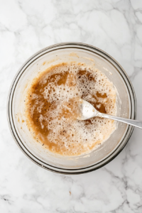 Top-down image of a clear glass bowl containing sautéed mushroom stems mixed with softened cream cheese, grated Parmesan cheese, black pepper, onion powder, and cayenne. A spoon is blending the ingredients into a smooth filling. This creamy, spicy mixture is used to stuff the mushroom caps before baking.