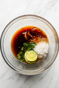 In this image, a clear glass bowl sits on a white marble countertop, filled with unwhisked dressing ingredients: rice vinegar, soy sauce, lime zest and juice, dark sesame oil, brown sugar, minced garlic, and red pepper flakes. A silver whisk lies next to the bowl. This tangy and flavorful dressing will coat the soba noodles for a delicious Asian-inspired salad.