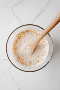 A top-down view of a transparent glass mixing bowl on a white marble countertop containing a mixture of warm water, flour, and active dry yeast. A wooden spoon with a light brown handle is partially submerged in the mixture, stirring it into a slightly bubbly consistency. This starter mixture is essential for activating the yeast and giving the bread a light and airy texture.