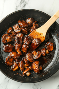 Mushrooms are now fully coated in a glossy brown oyster sauce mixture inside a black skillet set on a white marble countertop. The rich sauce blends with garlic, butter, and white wine to form a silky coating on the mushrooms. The dish looks deeply savory, creating a bold umami profile.