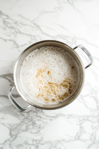 A top-down view of dry noodles being boiled in a large silver pot over a white marble countertop. The noodles are halfway cooked and visibly softening in bubbling hot water. This is the first step of preparing stir-fried soy garlic noodles. The clean and simple background focuses entirely on the pot and noodles in progress, showing the start of the cooking process clearly.