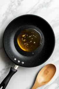 A clean top-down view of golden olive oil heating in a black nonstick pan over a white marble countertop. This step involves gently warming the oil to create a base for stir-frying the noodles, helping enhance the flavor and texture when combined with the savory teriyaki sauce.