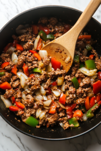A mixture of chopped onions, red bell peppers, green bell peppers, and minced garlic is being sautéed inside a black pot over a white marble countertop. The vegetables are softening in the flavorful sausage drippings. A wooden spoon stirs the colorful mixture, enhancing the dish’s aroma and texture.