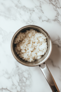 Top-down view of partially cooked white rice in a stainless steel saucepan, placed on a white marble countertop. The rice is simmering in water, with steam rising slightly. This image represents the initial preparation step for the stuffed cabbage rolls where the rice is pre-cooked until tender and ready to mix into the filling.