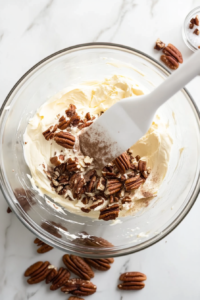 Top-down view of finely chopped pecans and kosher salt being stirred into the creamy cheese mixture in a clear glass bowl on a white marble countertop. A white silicone spatula is used for mixing. The pecans begin to blend into the smooth mixture, adding a nutty crunch that complements the sweetness of the other ingredients in the cheeseball.