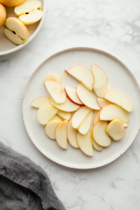 Peeled and cored apples are being sliced into thin, uniform pieces on a white plate. A stainless steel knife and vegetable peeler are visible on the side, placed neatly on a white marble countertop. This step highlights the importance of preparing sweet firm apples properly for even baking and aesthetic layering in the tart. Clean background and uniform slicing are emphasized in this essential prep stage.