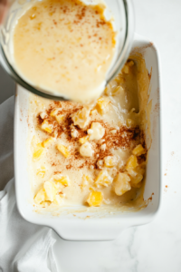 A top-down view of the smooth pineapple batter being poured from a clear glass bowl into a greased white 9-inch ceramic baking dish. The white marble countertop offers a clean, minimal background while the thick mixture with visible pineapple pieces is spread evenly, ready to go into the oven for baking.