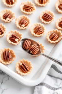 Pecan halves are being carefully placed into each of the mini pie shells that are arranged on a baking sheet. This image highlights the layering step before the sweet filling is added. Each shell gets a few pecans to provide texture, nutty flavor, and the signature look that defines classic pecan pie.