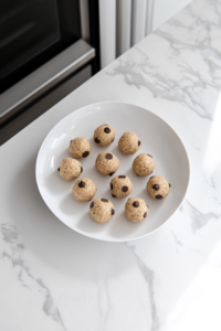 A white ceramic plate on a white marble surface holds several round, uniform energy bites lined up neatly. The chocolate chips and oats in each bite are visibly embedded, and the surface of each ball looks slightly moist and sticky. This step shows the preparation for chilling, with a focus on presentation and portion control.