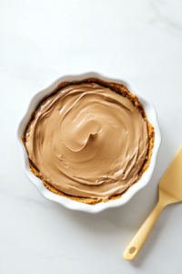 The chocolate cheesecake filling is poured and spread smoothly into the graham cracker crust inside a white ceramic 9-inch pie dish. A rubber spatula is used to level the surface. The dish rests on a white marble countertop, ready to be chilled. The rich, creamy texture is visibly smooth and well-blended.