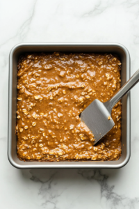 A top-down shot capturing the folding of finely diced candied ginger into the prepared ginger oat batter. The ginger pieces sit visibly atop the thick, dark mixture in a glass bowl, waiting to be gently incorporated. This optional step adds a sweet, spicy bite to every bar, enhancing the texture and flavor complexity.