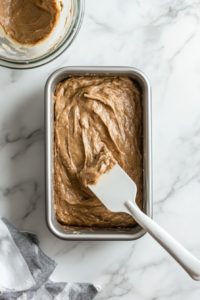 Top-down view of banana bread batter being poured into a greased metal loaf pan on a white marble countertop. A white spatula is used to smooth the top of the batter, ensuring even baking. The mixing bowl is set aside, now empty. The scene captures the final preparation step before placing the loaf into the oven for baking, keeping everything clean and visually uniform.