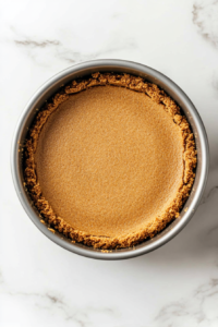 Top-down view of a round 8-inch oven-safe cake pan placed on a white marble countertop. The smooth cream cheese filling is being poured from the glass bowl onto the prepared graham cracker crust, filling the pan evenly. This step combines the two major layers, creating a base ready for air fryer baking.
