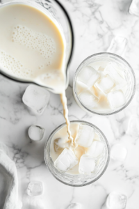 The creamy Brazilian lemonade is poured from a transparent glass blender into the ice-filled glasses. The drink flows smoothly over the ice, creating a cool, refreshing final presentation. Lime pulp and frothy texture are visible, showcasing the freshness of the ingredients. This step completes the recipe, ready for immediate enjoyment.