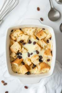 Top-down view of the whisked custard mixture being poured from a glass bowl over torn bread and raisins in a white ceramic baking dish. The bread begins to soak, absorbing the rich liquid. All ingredients and utensils are set over a clean white marble countertop, capturing the soaking step before baking.