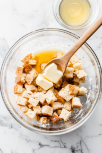A hand slowly pouring melted butter over the stuffing mixture while stirring with a wooden spoon. The bread cubes absorb the butter, ensuring the stuffing is moist, flavorful, and well-seasoned before baking.