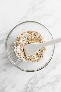 A stream of melted coconut oil is being poured into the bowl containing oats, flaxseed, cashew butter, and other dry ingredients. The white silicone spatula is mid-stir, working the oil evenly into the mixture. A small bowl with leftover oil and clean utensils are arranged on the white marble surface, capturing the moment of blending everything into a moist, workable dough.