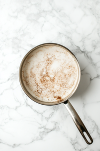 This top-down image captures the moment skim milk is being poured into a medium silver saucepan placed on a clean white marble countertop. The milk begins to fill the saucepan, indicating the start of this cozy beverage recipe. A glass measuring cup is nearby, adding a practical touch to the simple yet inviting kitchen scene.
