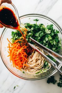 Pouring the dark, tangy soy sesame lime dressing into a large glass bowl containing soba noodles, grated carrots, and chopped cilantro. Salad tongs are placed beside the bowl on the white marble surface. This moment captures the merging of flavors as the noodles soak up the zesty dressing, bringing the Asian-style salad to life.