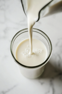 Top-down image showing 1 cup of unsweetened almond milk being poured into a clear glass blender jar on a clean white marble countertop. The blender sits empty except for the milk, with a measuring cup beside it. This image captures the start of the smoothie-making process in a clean, minimalist kitchen environment.