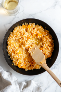 White wine is being poured from a glass measuring cup into the skillet with the rice and pumpkin mixture. This step deglazes the pan and infuses the risotto with depth and acidity. The image captures the moment before the wine is absorbed, with the remaining ingredients like stock and sage nearby, over a clean white countertop.