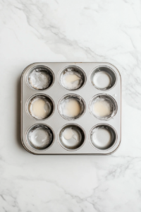 Top-down view of a clean silver muffin tin sprayed evenly with cooking spray, placed on a white marble countertop. The oven is preheated to 350°F, preparing for baking. This image captures the initial preparation step, where the muffin tin is primed to prevent sticking and ensure perfectly shaped mini quiches after baking.