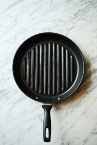 A clean black grill pan is preheating on a white marble cooktop in preparation for grilling cabbage steaks. The grill pan is empty but evenly heated, creating the ideal surface for crisping and charring vegetables. This step is essential to ensure the cabbage cooks evenly and develops those signature grilled flavors and textures that make this dish so delicious.