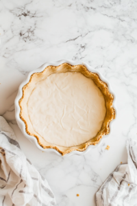 Top-down view of graham cracker crust being firmly pressed into a white 9-inch ceramic pie dish using a flat-bottomed glass. The crust forms an even base and sides, ready for chilling. The dish is placed on a bright white marble countertop, with a clean setup that highlights this crucial no-bake crust preparation step.