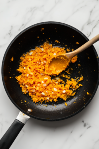 Top-down view of a black skillet sitting on a white marble countertop. The sautéed garlic and butter mixture is now combined with bright orange pumpkin puree, which is being stirred with a wooden spoon. A glass bowl with remaining pumpkin puree sits beside the skillet. This step blends the sweet earthiness of pumpkin with the aromatic garlic butter, creating a rich and flavorful base for the cream sauce.