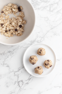 Hands are shown shaping the oat and chocolate chip mixture into 1-inch round energy bites. A few finished bites rest on a white ceramic plate, and the mixing bowl with leftover dough is visible in the background. The white marble countertop adds to the minimal, clean vibe, spotlighting the simple beauty of the homemade snack creation process.