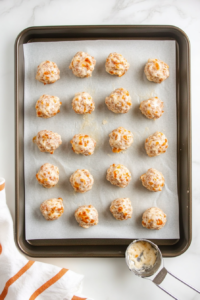 A parchment-lined baking sheet on a white marble countertop with small, evenly sized dough balls arranged in rows. The balls have a slightly rough texture, showing bits of cheddar cheese and sausage. A small metal scoop is placed beside the tray, indicating the portioning process for uniform size.