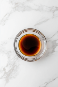 A small clear glass bowl filled with a dark and glossy sauce mixture made of soy sauce, teriyaki sauce, brown sugar, and mirin sits centered on a white marble countertop. Measuring spoons and small ingredients bowls surround it, making the setup clean and organized. This sauce will later coat the noodles with rich umami flavor and a hint of sweetness.
