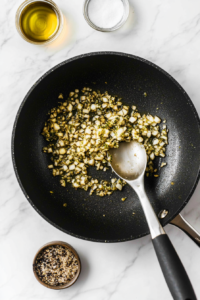 Top-down view of finely chopped mushroom stems being sautéed with minced garlic in hot vegetable oil inside a black skillet over a white marble countertop. The garlic is golden and aromatic, blending with the mushrooms for a rich base. This step brings out deep flavors for the creamy stuffing.