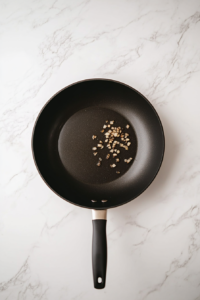Top-down view of a black skillet on a white marble countertop, where diced onions and minced garlic are being sautéed in melted unsalted butter. The ingredients are sizzling gently, releasing a fragrant aroma that forms the flavor base of the pumpkin risotto. A wooden spatula stirs the mixture while glass bowls of prepped ingredients sit nearby.