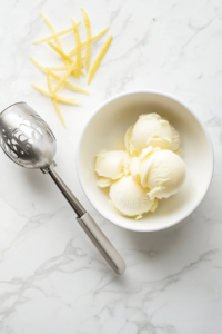 A stainless steel ice cream scoop is lifting pale yellow lemon sorbet from a glass container, and placing it into a simple white ceramic bowl. The frozen sorbet is garnished with delicate lemon zest strips, making it visually refreshing and vibrant. The entire scene is styled on a clean white marble countertop.