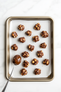 A spoon is being used to scoop small mounds of the chocolate and peanut mixture onto a baking sheet lined with parchment paper. Each cluster is carefully placed to avoid sticking, and the melted chocolate glistens under the light. The tray rests on a clean white marble surface, ready for cooling.