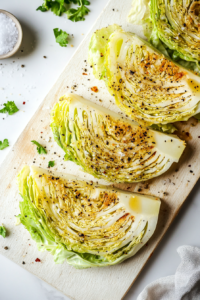 Cabbage steaks, already brushed with olive oil and garlic, are being seasoned with salt and freshly ground black pepper on a white cutting board placed over a white marble surface. Small bowls of salt and pepper are visible, emphasizing the simplicity and freshness of the seasoning. This final prep step is essential for enhancing the natural flavors before grilling.