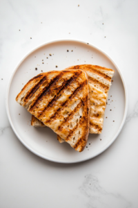 A grilled four-cheese sandwich is served warm on a white ceramic plate with a small bowl of tomato soup beside it, placed on a white marble countertop. The sandwich is sliced diagonally, showing off the layers of melty cheese. This final image captures the enjoyment of the meal, suggesting pairing ideas and presenting the dish as comforting and delicious. The scene is cozy, inviting, and ideal for serving suggestions.
