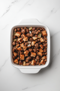 A serving spoon scooping warm, freshly baked sausage stuffing from the casserole dish. The stuffing has a perfect combination of crispy golden top and moist, flavorful inside, making it a delicious and comforting side dish for any meal.