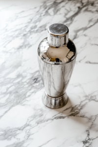 This image shows the third step of the Mistletoe Martini recipe, where the cocktail shaker is being held and shaken vigorously over a white marble countertop. Inside the shaker are vodka, cherry brandy, lime juice, simple syrup, and ice. The action shot captures the energy and motion needed to properly blend and chill the ingredients. This step is essential for a well-balanced martini and ensures the flavors are fully incorporated. The consistent color theme and minimalist setup add elegance and clarity to the visual instructions.
